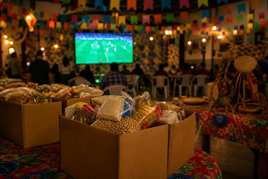 Caixas de alimentos em festa junina comunitária com telão de futebol ao fundo, usadas como capa sobre o Arraial Solidário